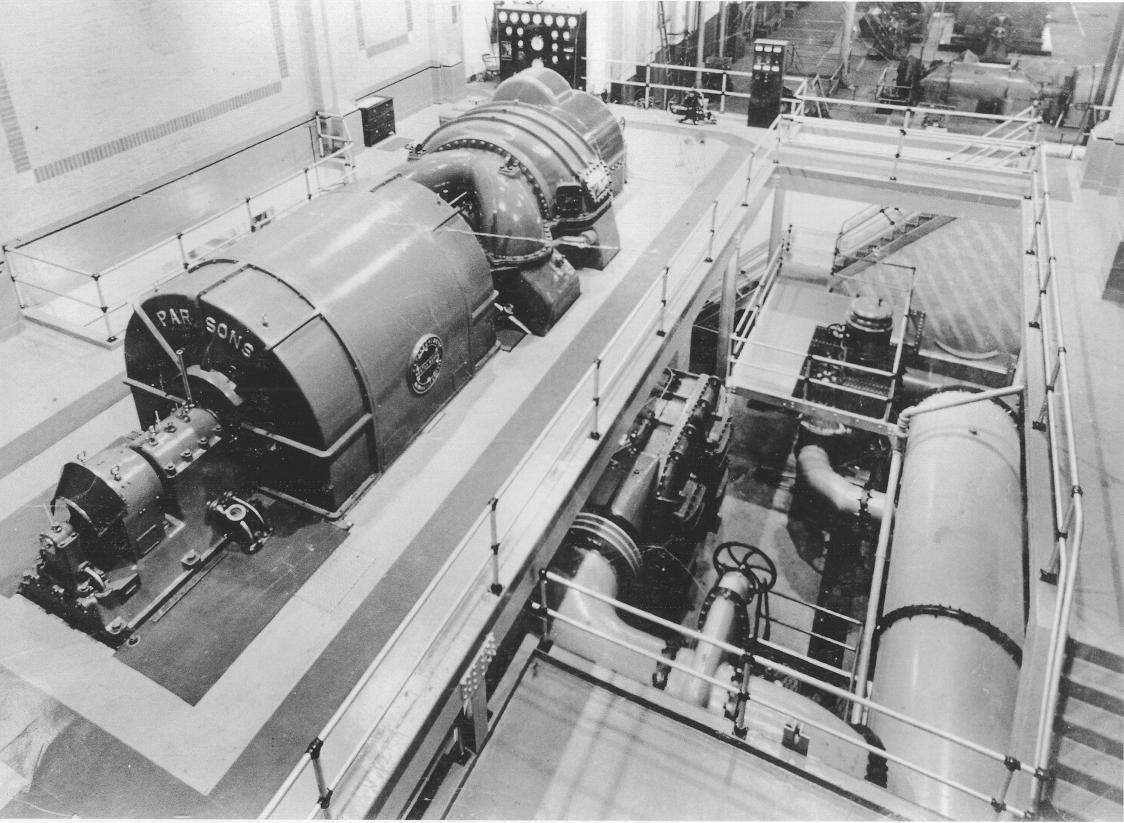 Parsons #1 with generator/exciter in foreground and turbine and control panel in background. Cooling water lines in lower level to the right of generating unit.